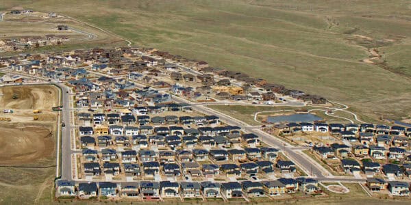 A newly built suburban neighborhood: rows of similar detached houses sit on gridded streets, with nearby construction sites, a small pond and walking paths, bordered by open grassy fields.