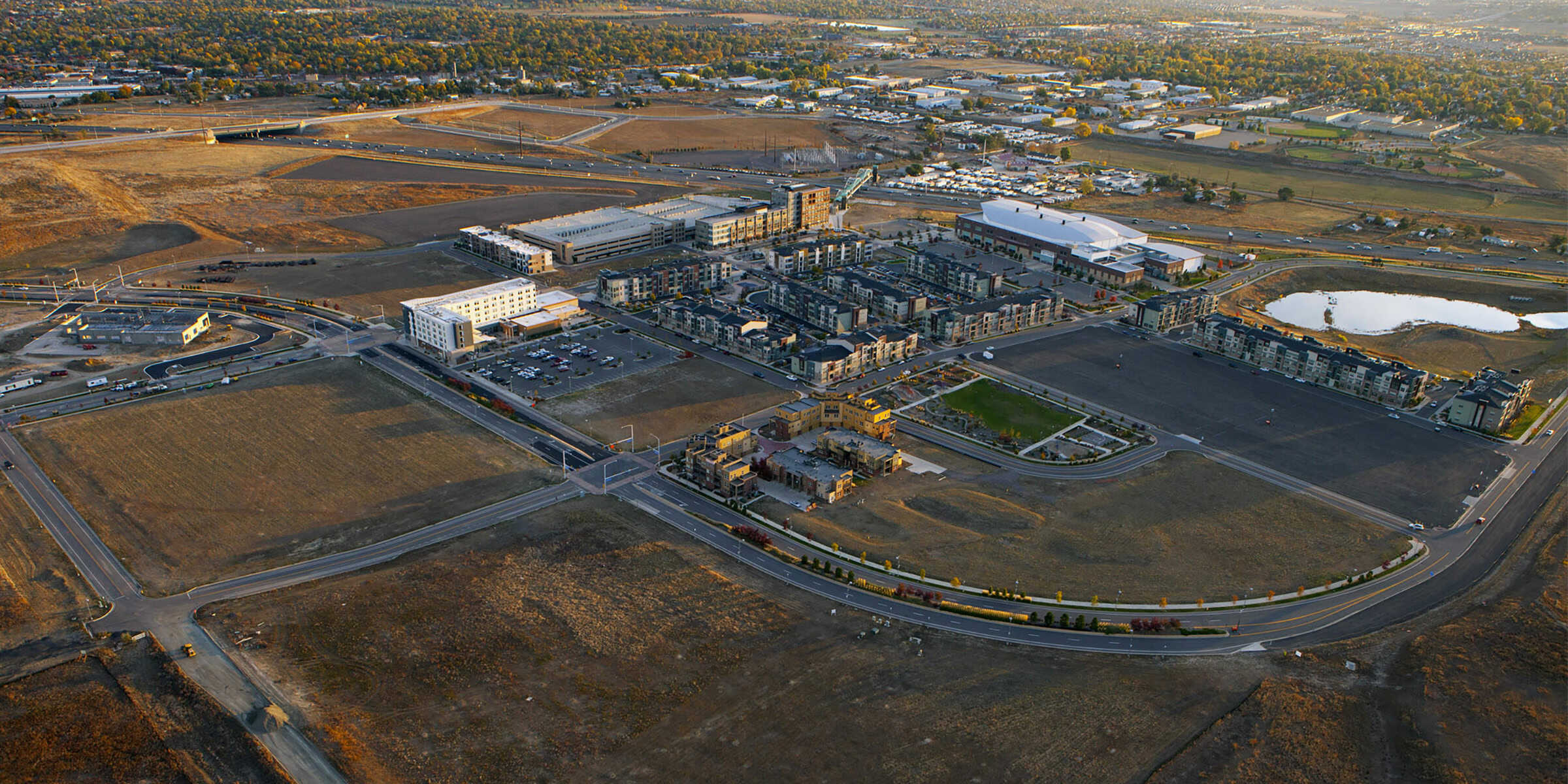 A cluster of mid-rise apartment buildings sits amid developing parcels, surrounded by curved roads, empty brown fields, a small pond, and distant residential and industrial areas at sunset.