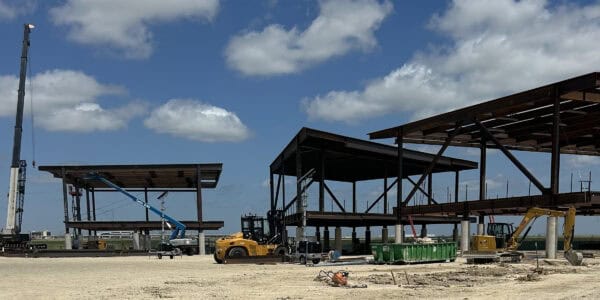 Steel building frames rise, being erected by cranes and lifts amid construction machinery, standing on concrete piers at a flat, dusty site under a blue, partly cloudy sky.

Visible text: "Genie S-85" on blue boom lift; "CAT" on yellow excavator.