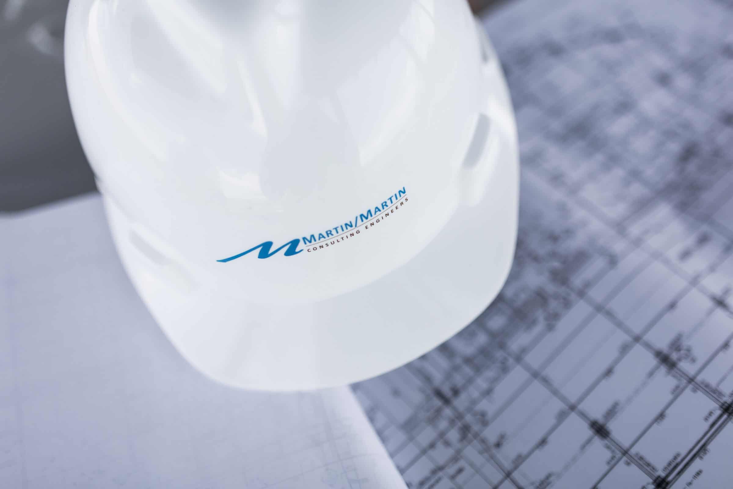 White hard hat sits on architectural blueprints on a table, close-up. Text on helmet: "MARTIN/MARTIN CONSULTING ENGINEERS".