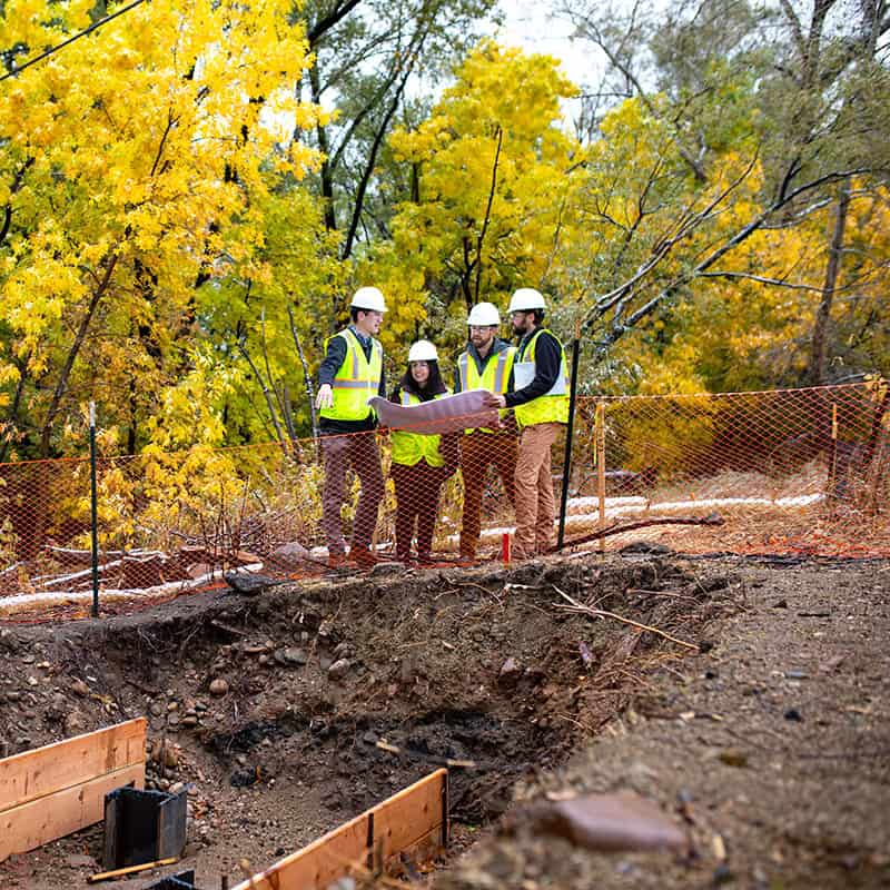Four construction workers wearing hard hats and high-visibility vests examine blueprints beside an orange safety fence at a forested excavation site with yellow autumn trees in the background.