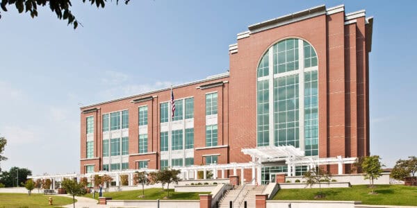 Four-story red-brick civic building stands with tall arched glass window, flanked by rows of rectangular windows; steps, white pergola and American flag on landscaped lawn under a clear blue sky.