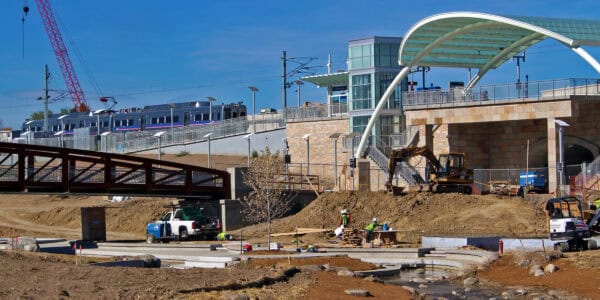 Excavator digs earth while construction workers move materials near a partially built transit station and elevated train platform; trucks, a pedestrian bridge and clear blue sky surround the dusty site.