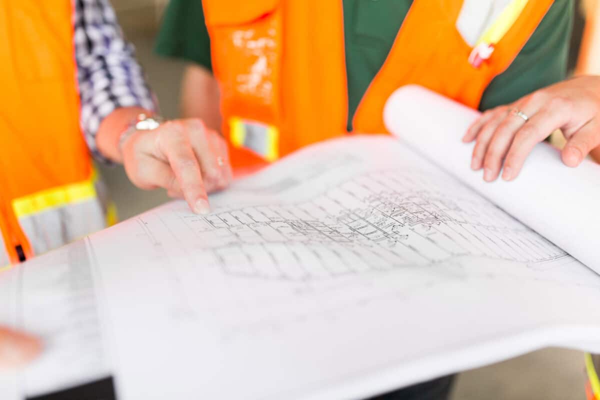 Architectural blueprint spread on a table, two people point to and hold its pages, wearing orange safety vests and casual shirts in a construction-site or indoor work-area setting.