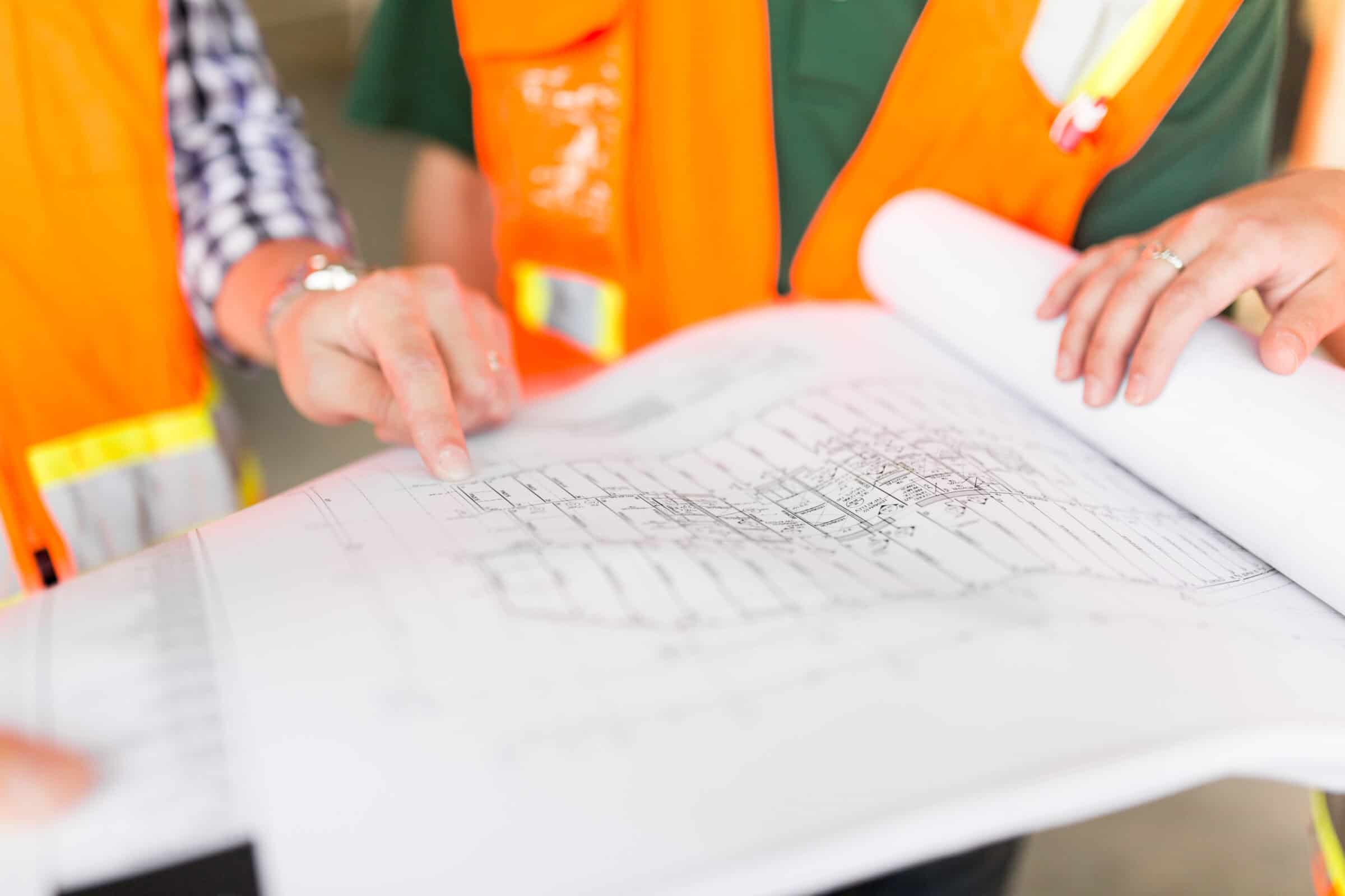 Architectural blueprint spread on a table, two people point to and hold its pages, wearing orange safety vests and casual shirts in a construction-site or indoor work-area setting.