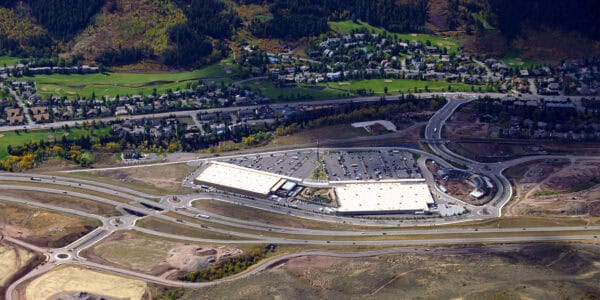 Large retail complex occupies land between a multilane highway and a suburban neighborhood with golf-course greens; expansive parking lots, multiple roundabouts and ramps connect to surrounding hills and forested slopes.