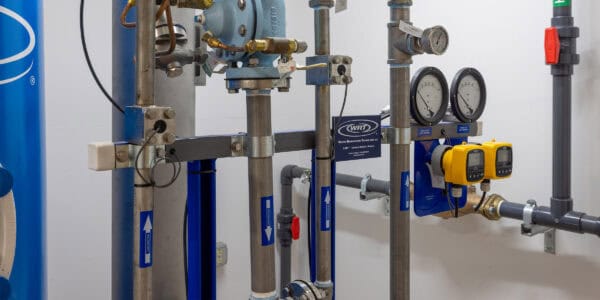 Stainless-steel pipes and gauges form a plumbing assembly, measuring flow and pressure, mounted on a white mechanical-room wall with blue tanks and two yellow digital meters.

Visible text: "WRT" ; "Water Remediation Technology LLC"