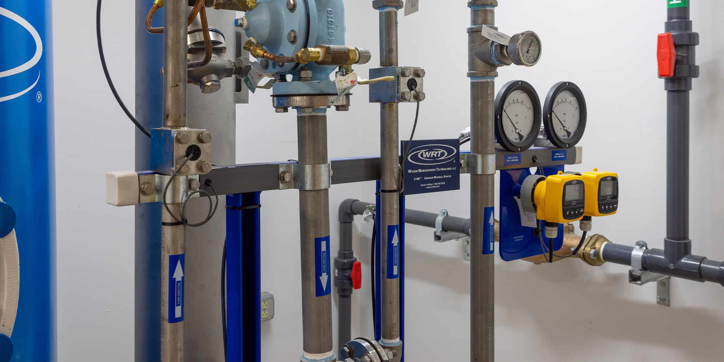Stainless-steel pipes and gauges form a plumbing assembly, measuring flow and pressure, mounted on a white mechanical-room wall with blue tanks and two yellow digital meters.

Visible text: "WRT" ; "Water Remediation Technology LLC"