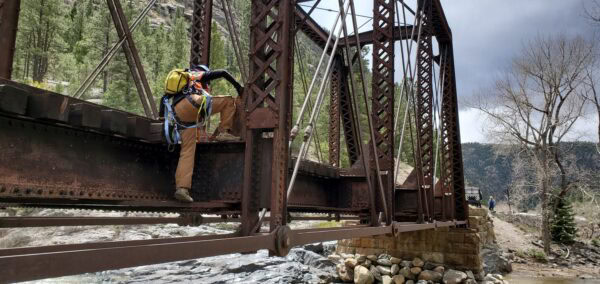 Worker in a yellow backpack and safety harness climbs a rusted iron truss bridge girder above a rocky river, framed by pine-covered canyon slopes and a cloudy sky; another worker stands on the far bank.