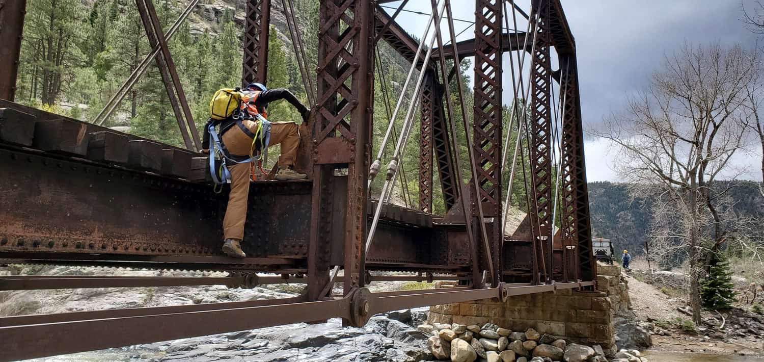 Worker in a yellow backpack and safety harness climbs a rusted iron truss bridge girder above a rocky river, framed by pine-covered canyon slopes and a cloudy sky; another worker stands on the far bank.