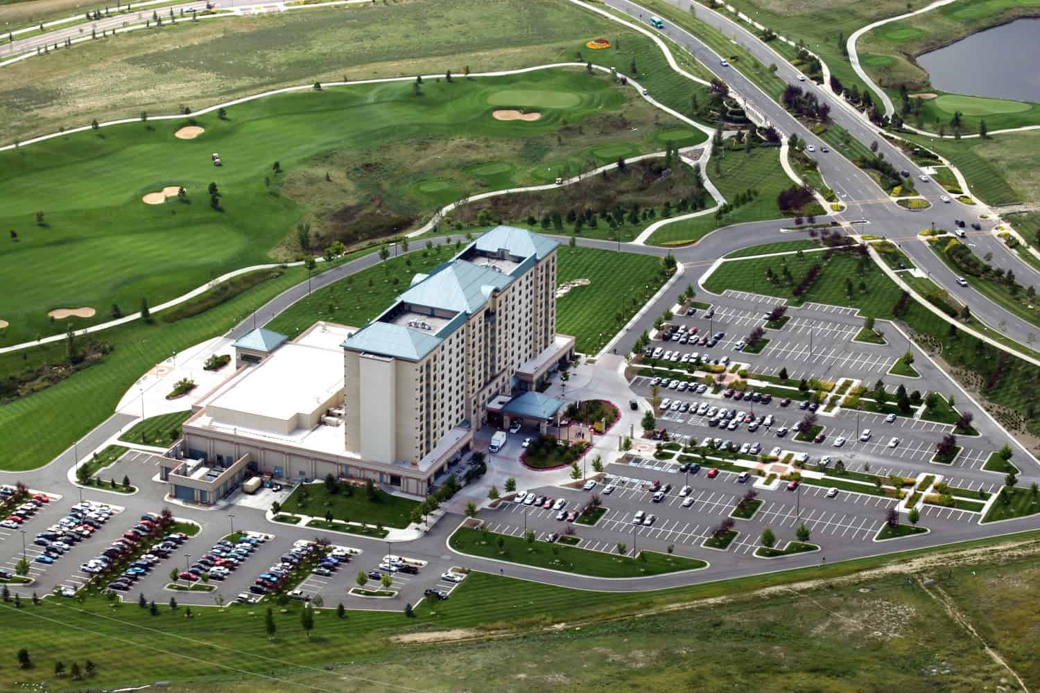 Multi-story hotel complex anchors a landscaped campus, surrounded by dozens of parked cars in extensive parking lots, adjacent to manicured golf-course fairways, winding paths, and intersecting roads.