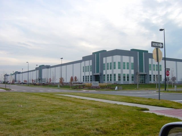 Large industrial warehouse stretches along a suburban road; grassy foreground and sidewalks; street signs read "ONE WAY" and "STOP"; overcast sky.