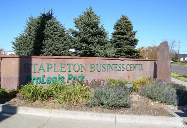 Stone sign reads "STAPLETON BUSINESS CENTER" and "ProLogis Park", standing amid landscaped beds and evergreen trees beside a roadway under a clear blue sky.