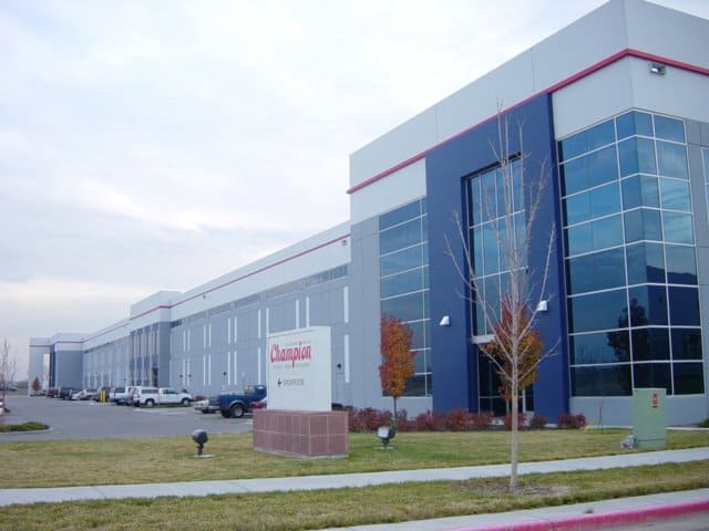 Large gray industrial warehouse building stretches back; sign reads "Champion". Several parked trucks sit near loading bays; small trees and a grassy lawn line the street.