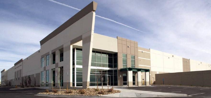 Modern beige warehouse-office building anchors the scene, with tall glass windows and a green-awned entrance, bordered by empty parking, rock landscaping, and a clear sky streaked by a contrail.