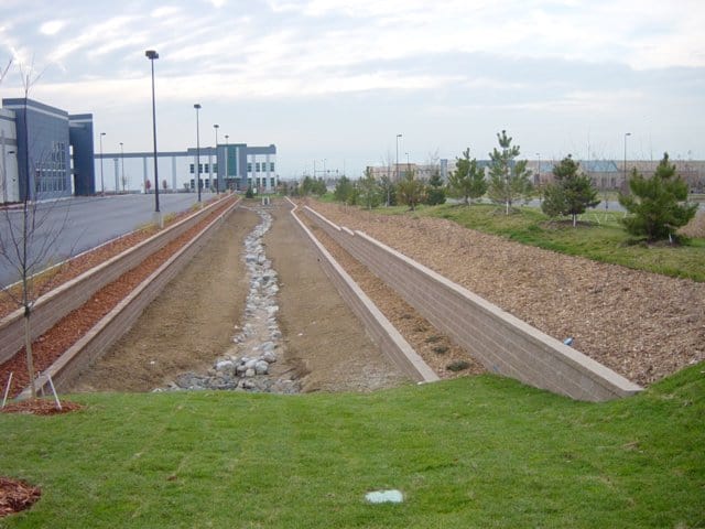 Dry concrete-lined drainage channel with a rocky bed runs toward distant low-rise industrial buildings, flanked by grassy slopes, mulch beds, young trees and lamp posts under a cloudy sky.