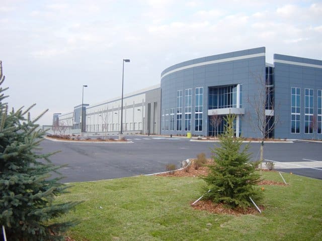 Large modern industrial warehouse sits behind an empty asphalt parking lot, flanked by young evergreen trees and landscaped grass, under an overcast sky with tall lamp posts along the building.