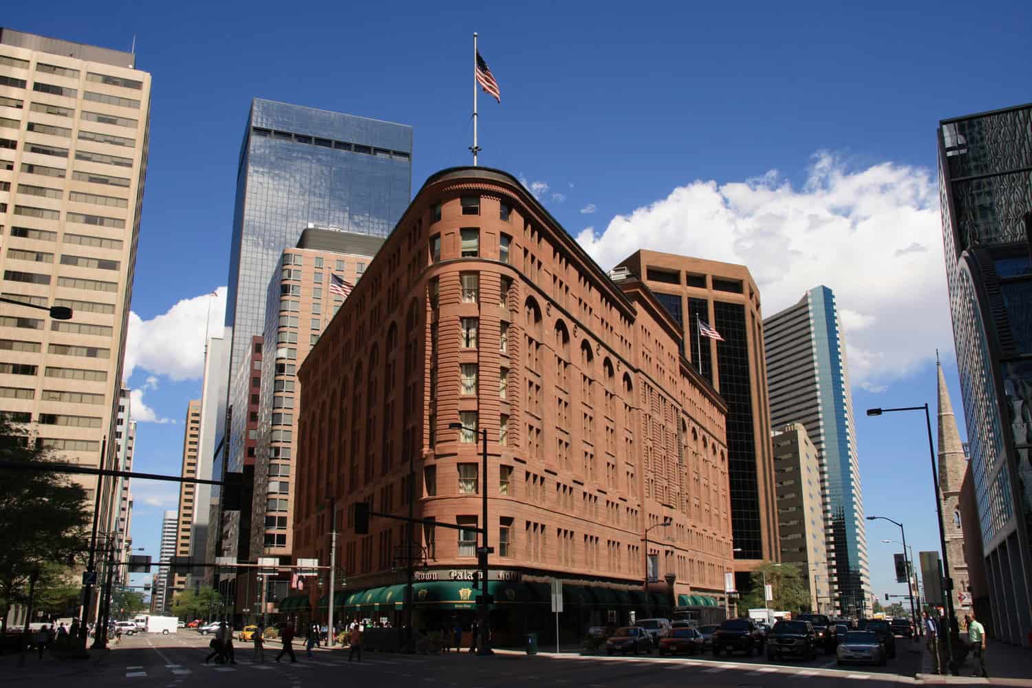 Red-brown flatiron hotel dominates the intersection, flying an American flag; pedestrians cross and cars queue along a downtown street flanked by glass skyscrapers under a bright blue sky.

Visible text on awning: "Brown Palace Hotel"