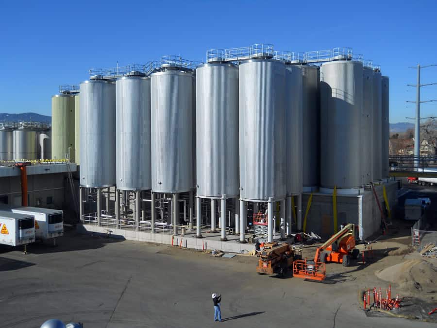 Large cylindrical metal storage silos stand on a concrete platform, clustered together as construction equipment and a lone hard-hat worker occupy the industrial yard beneath a clear blue sky.