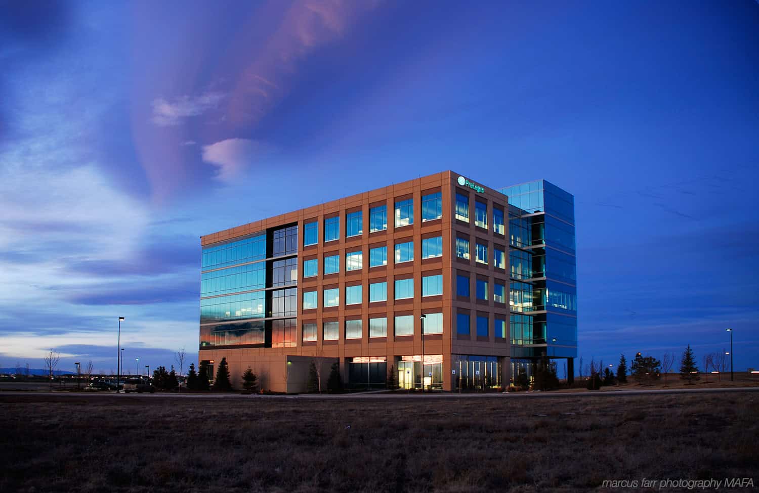 A glass office building reflects sunset, its lit windows glowing; it stands on a grassy plain under a deep blue dusk sky with sweeping clouds. Visible text: marcus farr photography MAFA