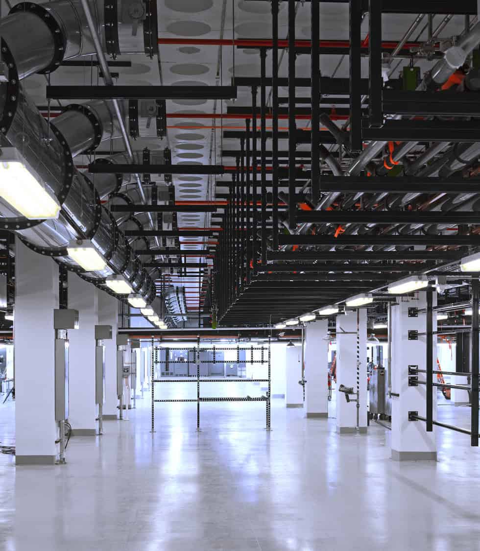 Overhead metal pipes and black cable trays span the ceiling, supported by white columns, illuminating an empty polished-concrete industrial utility space with suspended fluorescent lights.