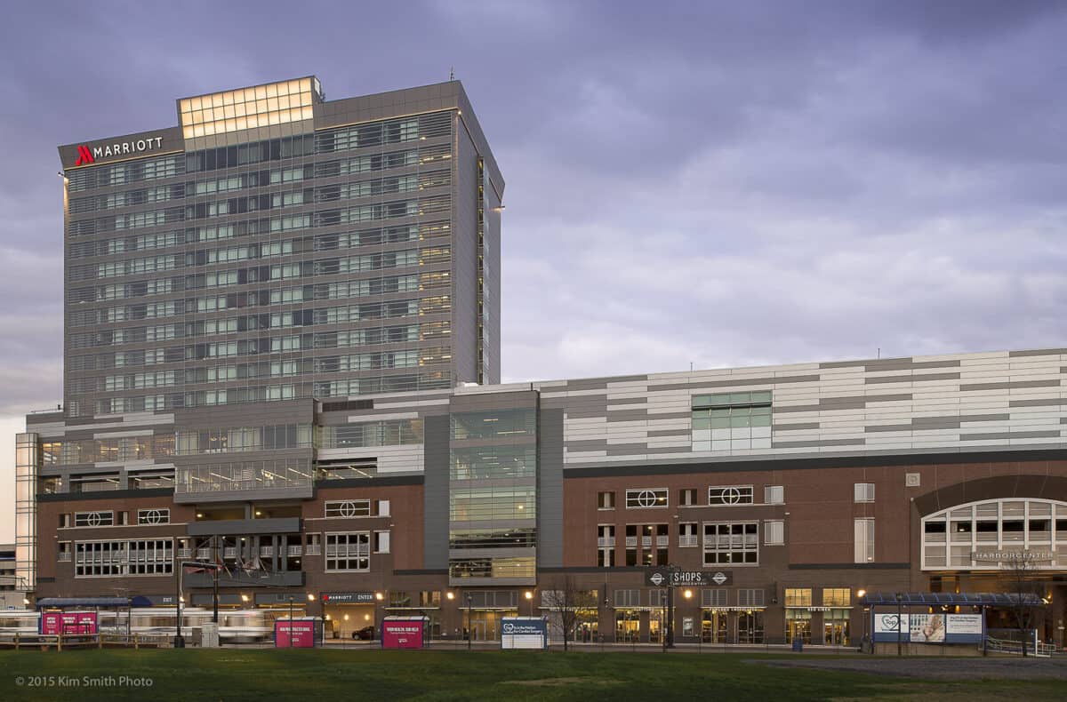 Marriott hotel tower rises above a multi-story brick-and-glass retail complex, facing a grassy foreground under a broad cloudy sky.

Transcribed text: MARRIOTT; SHOPS; HARBORCENTER; © 2015 Kim Smith Photo