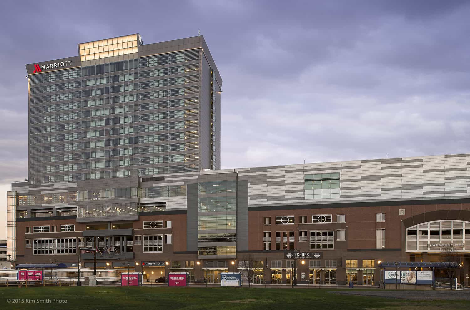 Marriott hotel tower rises above a multi-story brick-and-glass retail complex, facing a grassy foreground under a broad cloudy sky.

Transcribed text: MARRIOTT; SHOPS; HARBORCENTER; © 2015 Kim Smith Photo