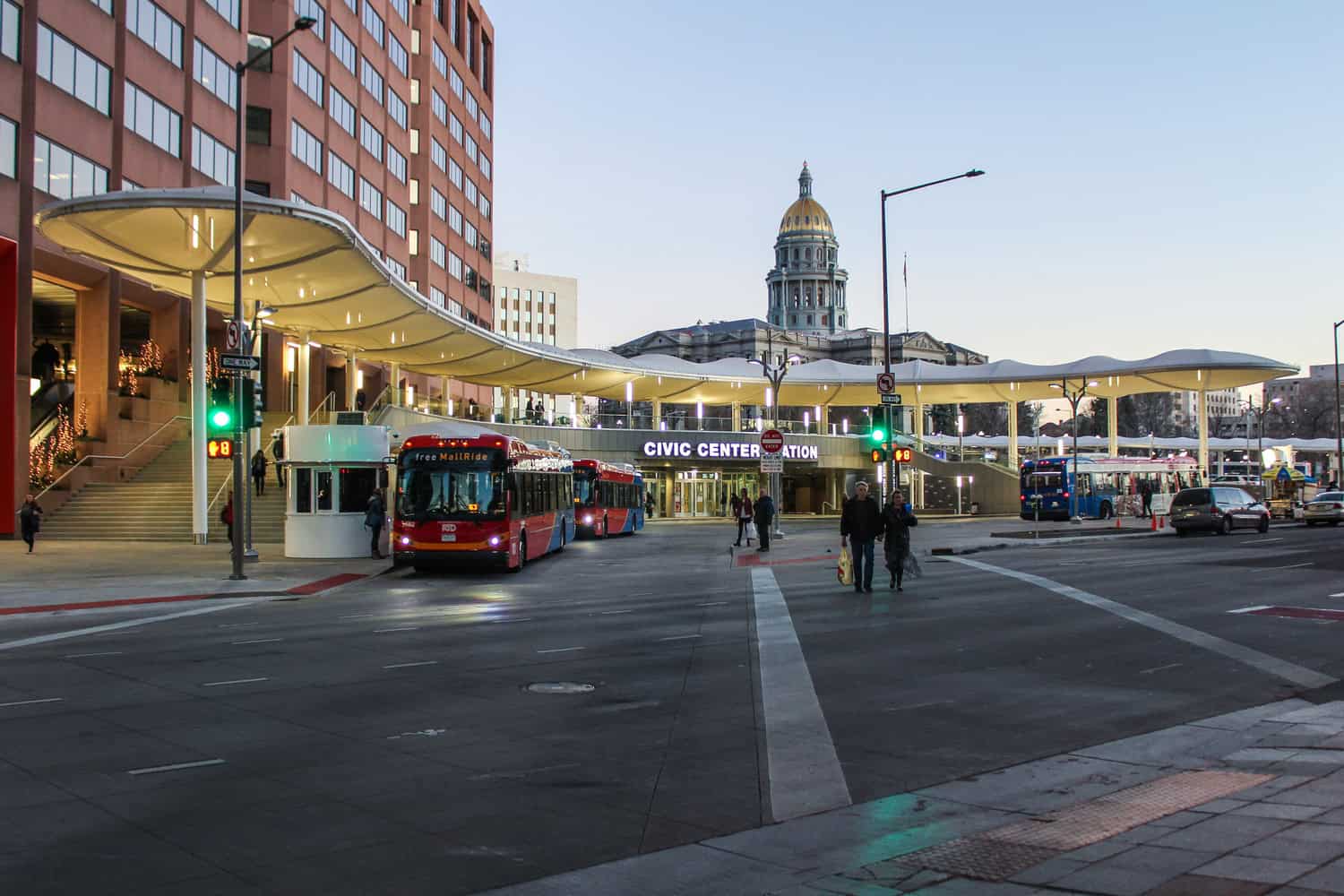 A covered bus-station canopy shelters red and blue buses while passengers cross a downtown intersection, the golden-domed capitol building rising behind the Civic Center at dusk.

Text: "CIVIC CENTER STATION", "free MallRide", "ONE WAY"