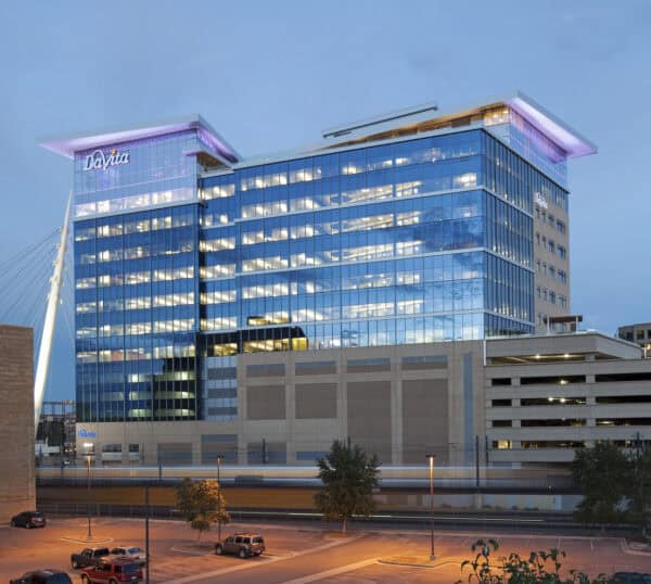 Glass office tower (sign reads "DaVita") glows with interior lights, housing active offices; street-level light rail blurs past and a lit parking lot sits in front under a dusky sky.