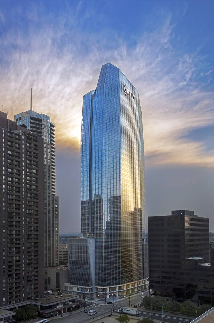 A glass-clad skyscraper reflects the golden sunset, rising above a city intersection while neighboring high-rises frame it and streets bustle below. Text on building: "Sheraton".