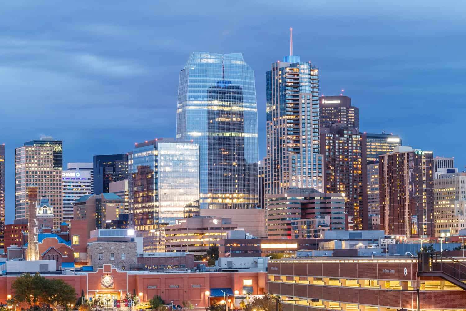 A glass skyscraper reflects interior lights while towering over surrounding high-rises and a low brick parking garage at dusk beneath a broad, cloudy blue sky.

Visible text: "CHASE"; "TIVOLI"; "7th Street Garage"