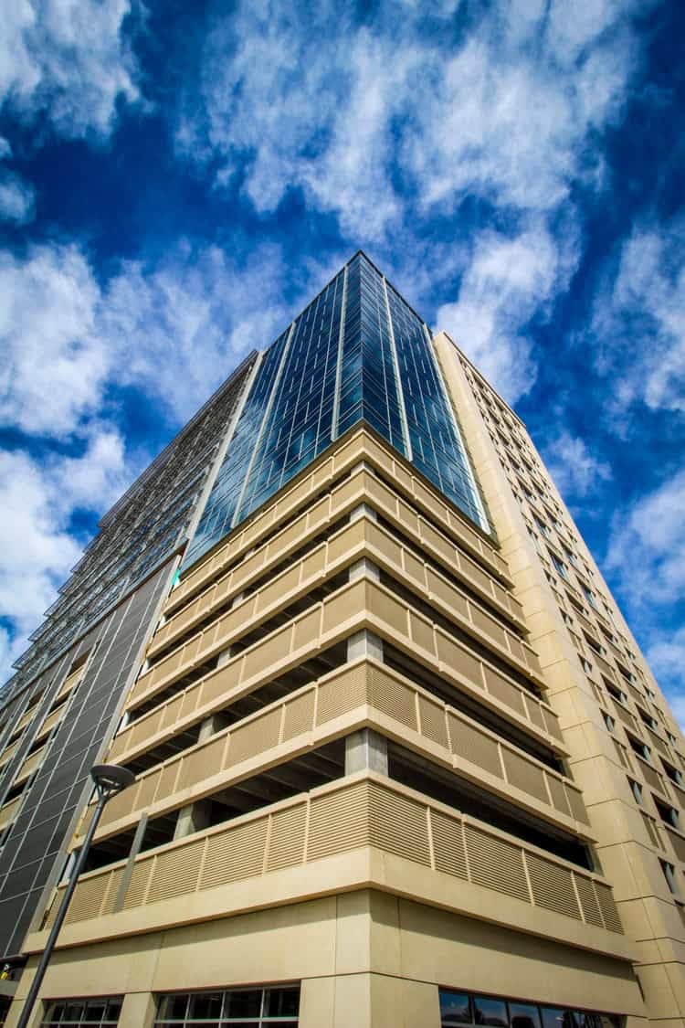 High-rise building rises from a low-angle view, its beige louvered parking base supporting reflective glass upper floors, set against a vivid blue sky streaked with white clouds.