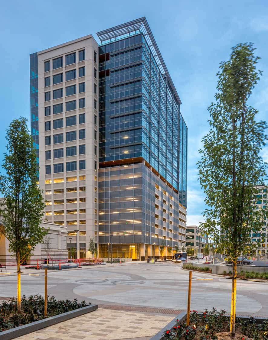 Glass-and-concrete office tower rises above a paved plaza, its lit windows reflecting dusk. Young trees border the open square; surrounding modern buildings and soft evening sky complete the urban scene.