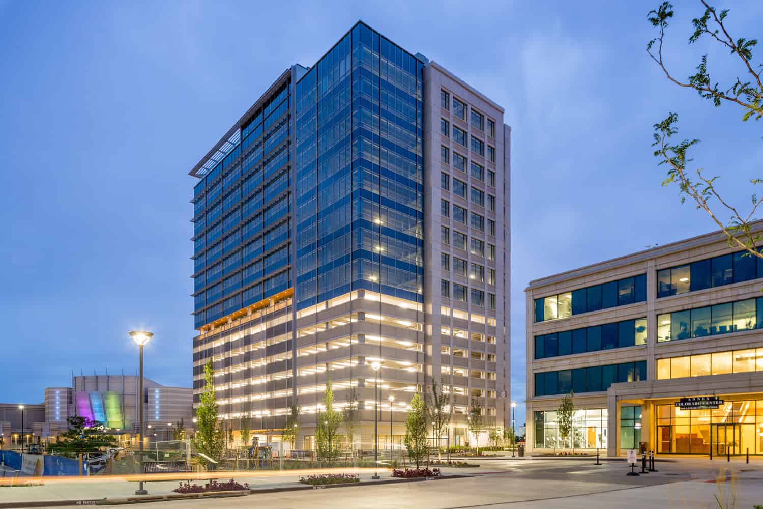 Glass-and-concrete office tower rises, interior lights on, flanked by lower office buildings and streetlamps in a dusk urban plaza.

Transcribed text: "ANNEX COLORADO CENTER" (entrance). Curb stencil: "NO PARKING".