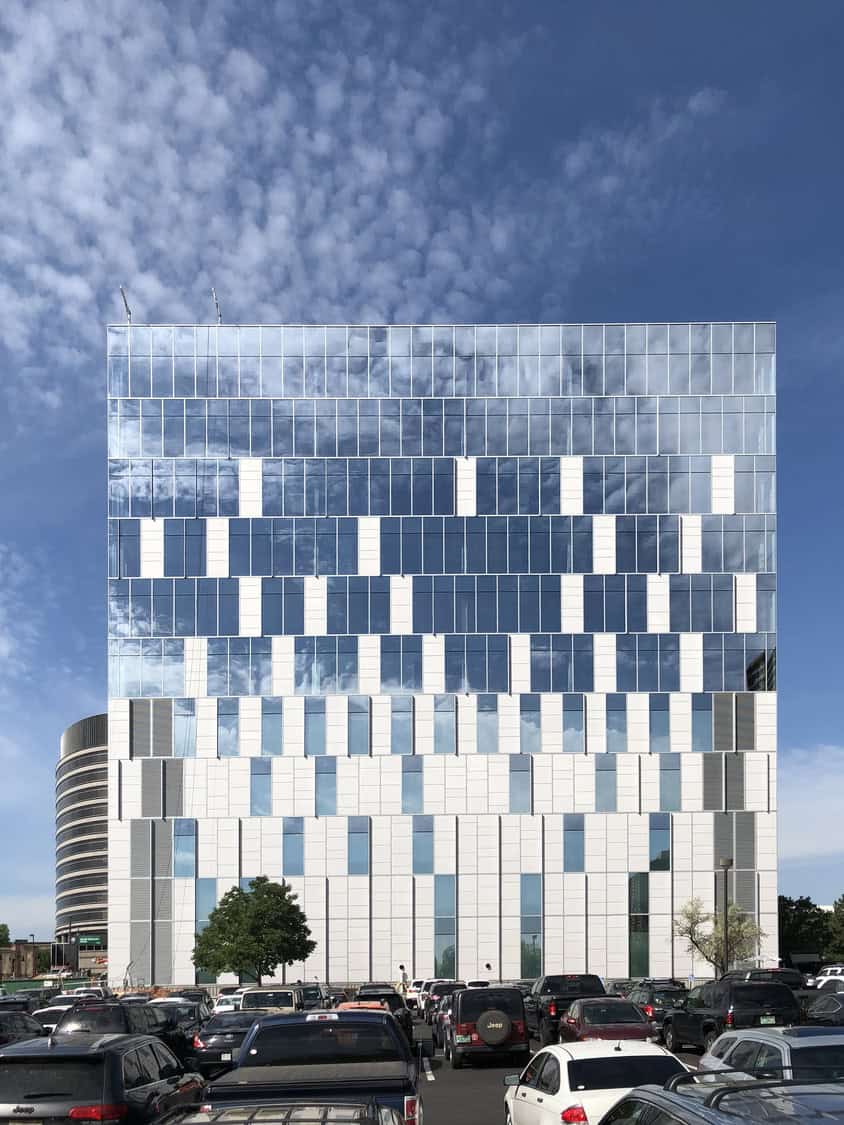 A modern glass-and-white-paneled office tower reflects clouds and blue sky, rising behind a crowded parking lot of cars and SUVs beneath a bright, partly cloudy sky.