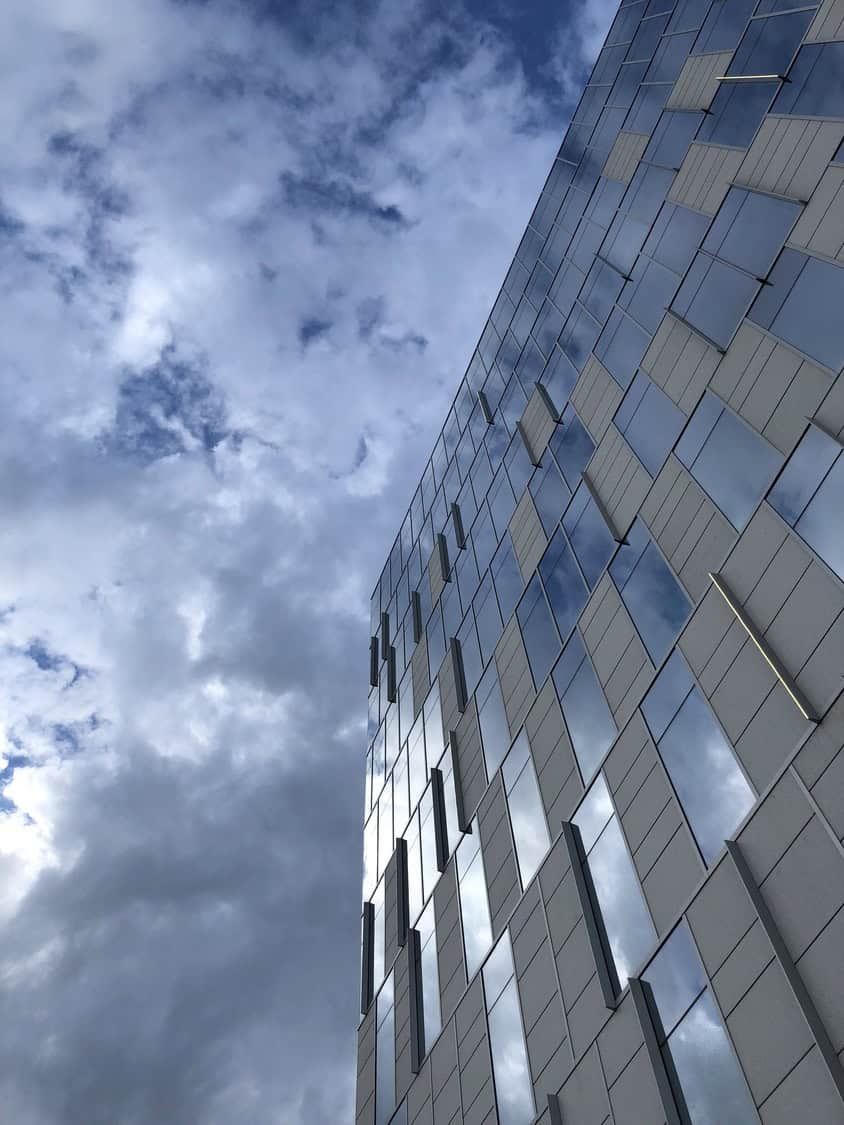 A glass-faced modern building rises diagonally, its rectangular windows and vertical fins reflecting brooding clouds, set against an expansive overcast sky with varied gray-blue cloud formations.