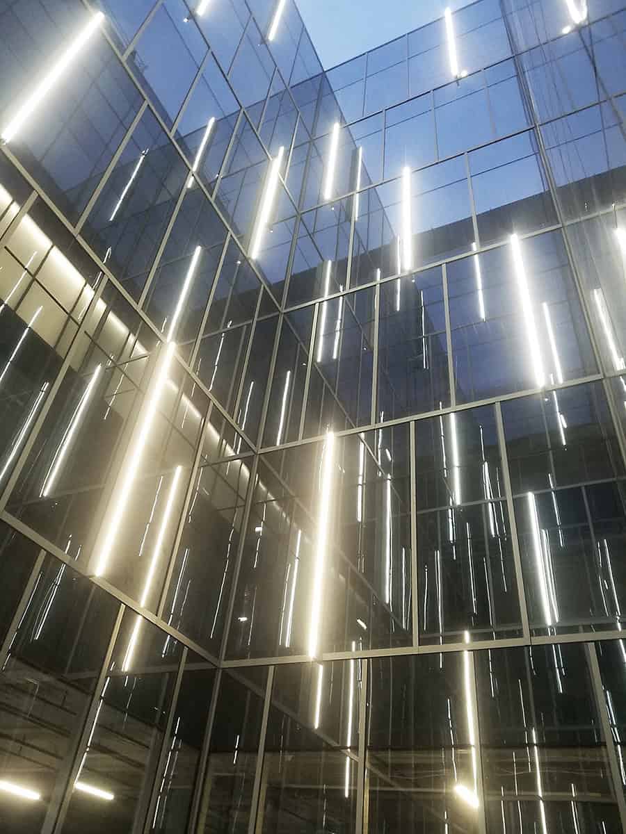 Glass atrium wall adorned with vertical LED light tubes, their reflections multiplying across a grid of floor-to-ceiling windows in a modern building courtyard beneath a dusky blue sky.