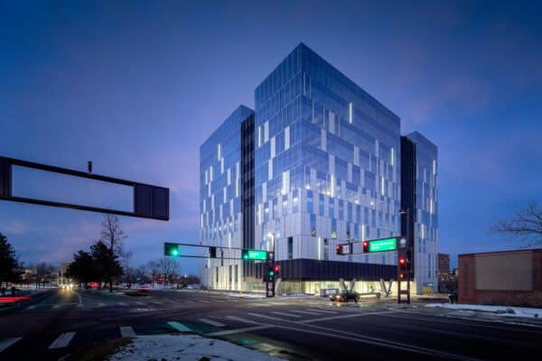 A glass office building glows with interior lights, standing at a snowy urban intersection at dusk while traffic lights and a few cars move past under a deep blue sky.

Transcription: "South Syracuse Street" (other overhead sign text illegible).