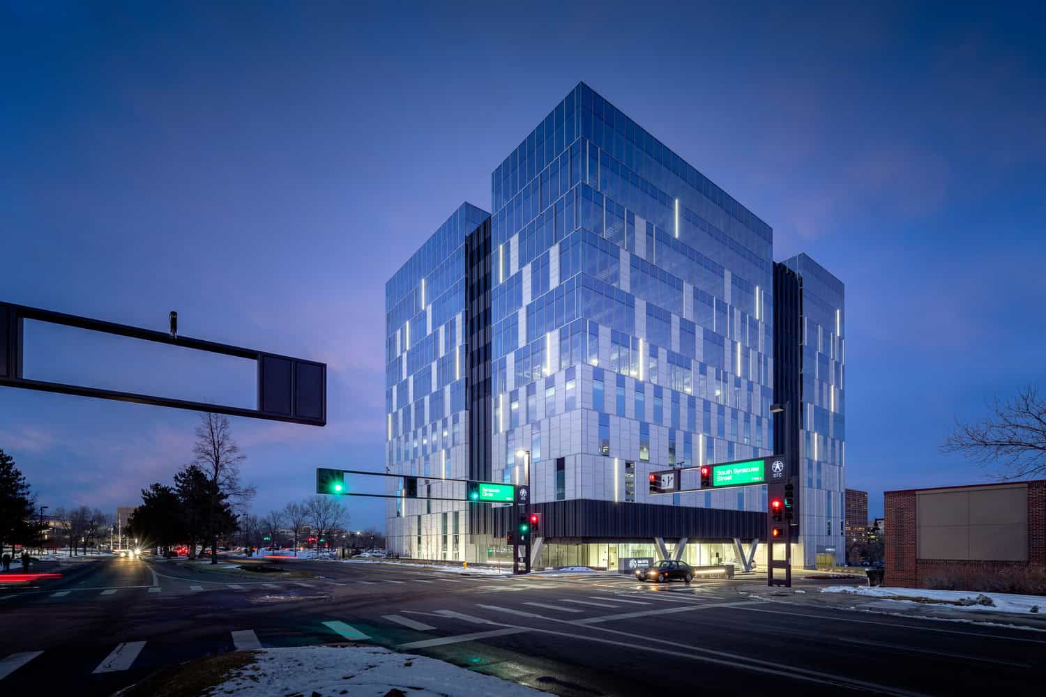 A glass office building glows with interior lights, standing at a snowy urban intersection at dusk while traffic lights and a few cars move past under a deep blue sky.

Transcription: "South Syracuse Street" (other overhead sign text illegible).
