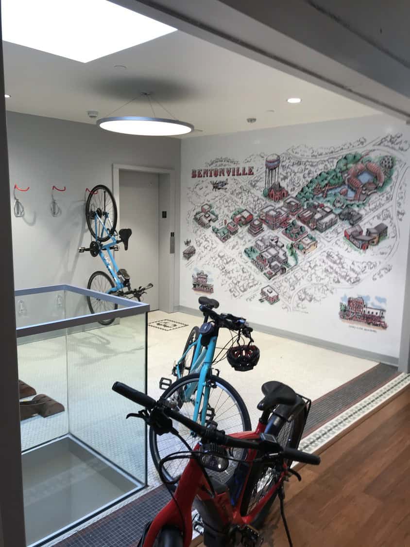 Bicycles parked and stored: one hung vertically on a wall rack, two standing by a glass stairwell; wall features a detailed town map mural. Text visible: "BENTONVILLE".