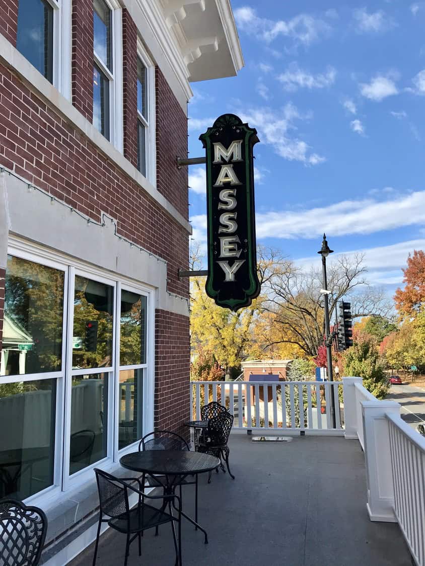 Vertical sign reading "MASSEY" hangs from a brick building; metal bistro tables and chairs occupy a second-floor balcony overlooking a street lined with autumn trees under a blue sky.