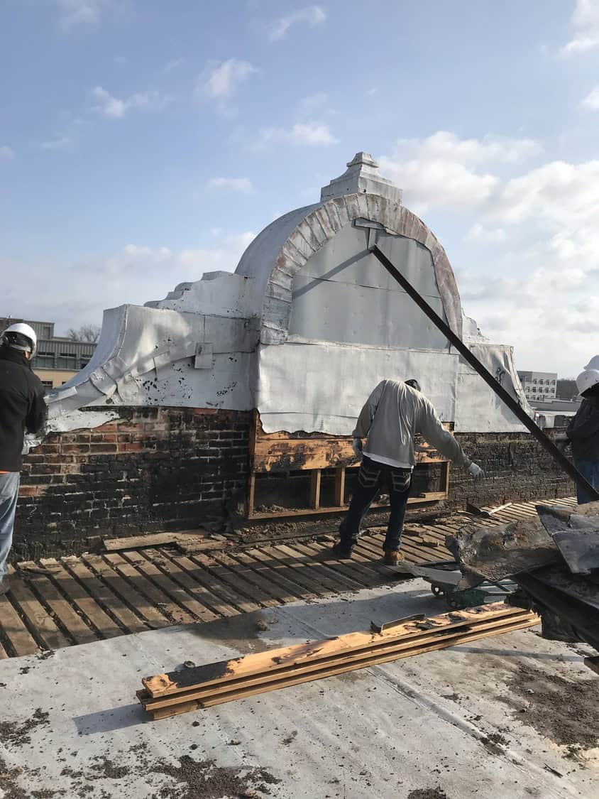 White curved rooftop parapet being stripped by construction workers, exposing brick and wooden framing on a flat roof with scattered planks and tools beneath a partly cloudy sky.