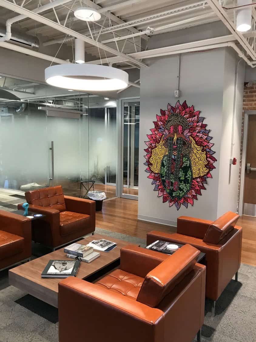 Colorful mandala-style mural adorns a wall; four orange leather armchairs face a low wooden coffee table with books, set in a glass-walled, modern office lounge under a circular ceiling light.