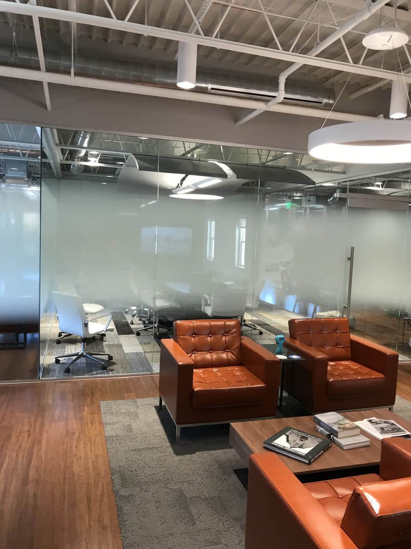 Two tufted orange leather armchairs sit beside a low wooden coffee table, arranged in a modern office lounge with frosted glass-walled conference room, exposed ductwork, and hardwood flooring.