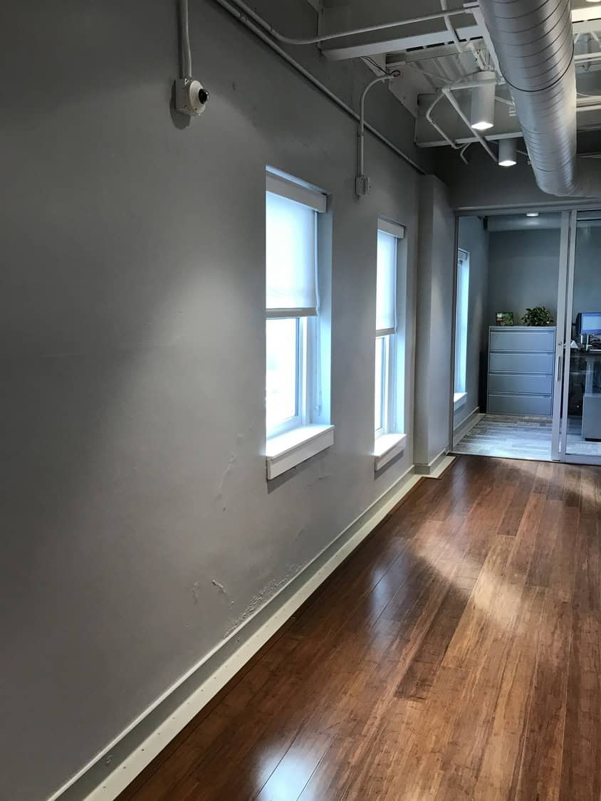 Narrow office corridor with polished wooden floor; sunlight streams through three recessed windows, leading to a glass-walled workspace with filing cabinet; exposed ceiling ducts and a wall-mounted security camera.