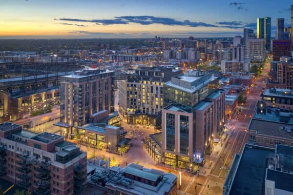 A cluster of modern mid-rise buildings glows with interior lights, framing a central pedestrian plaza while streets stretch through a twilight downtown toward a distant skyline and setting sun.