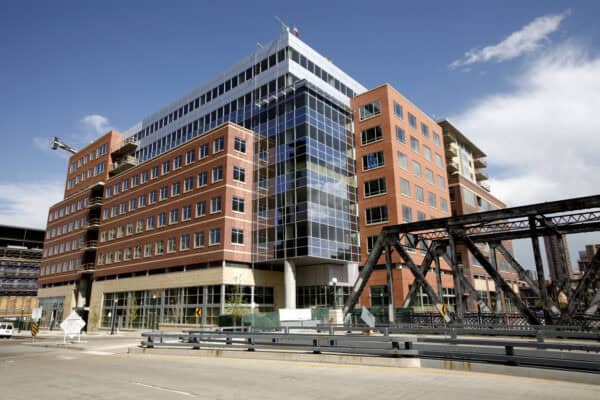 Multi-story brick-and-glass office building rises, its glass corner reflecting clouds; an aged steel truss bridge crosses the street in a downtown urban setting under a blue sky.