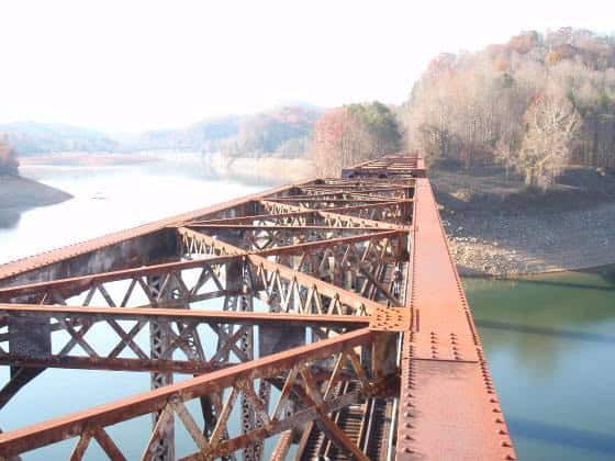 Rusty steel railway trestle stretches across a calm reservoir toward tree-covered hills with autumn foliage, its lattice reflected in the water beneath a pale sky.