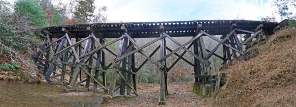Wooden railroad trestle spans a shallow creek, its angled timber supports crisscrossing beneath a track deck, set amid bare trees and brown, leaf-strewn banks in a quiet forest.