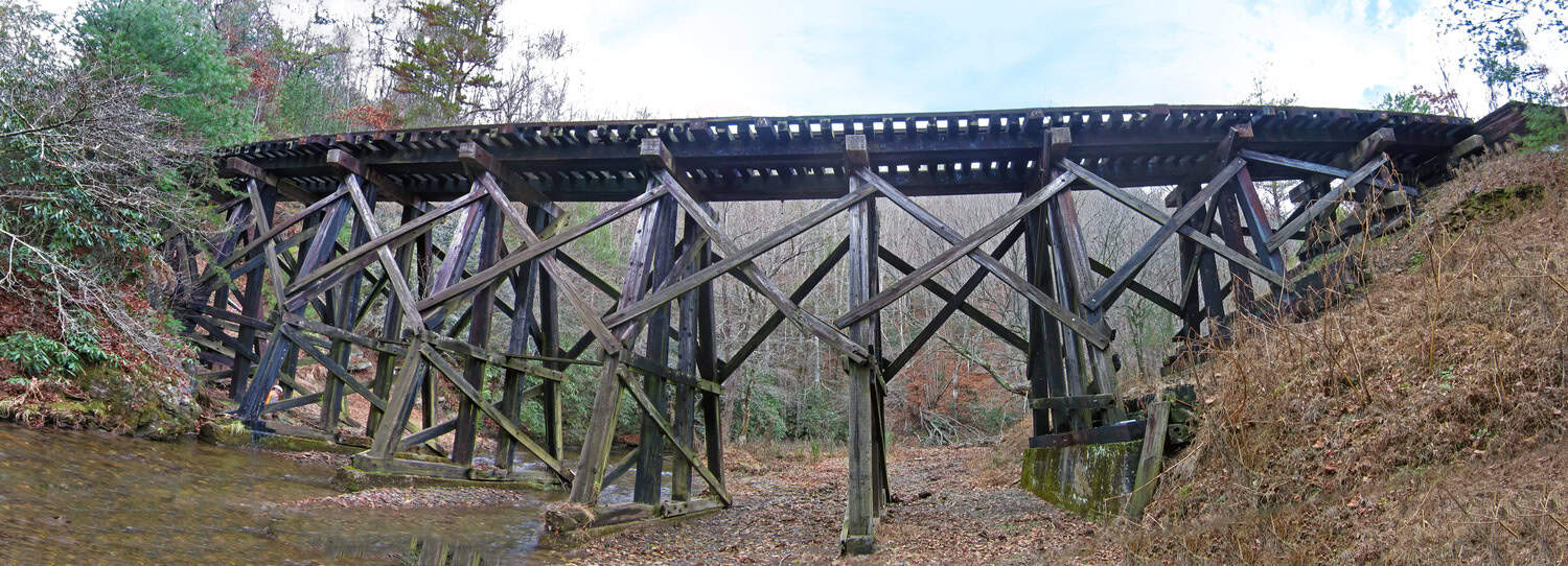Wooden railroad trestle spans a shallow creek, its angled timber supports crisscrossing beneath a track deck, set amid bare trees and brown, leaf-strewn banks in a quiet forest.